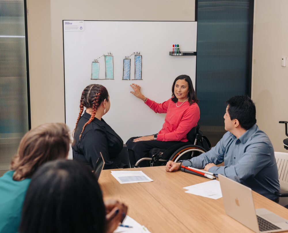 A woman using a wheelchair presents data to her colleagues during a meeting. She gestures to illustrated visualizations on a whiteboard behind her.
