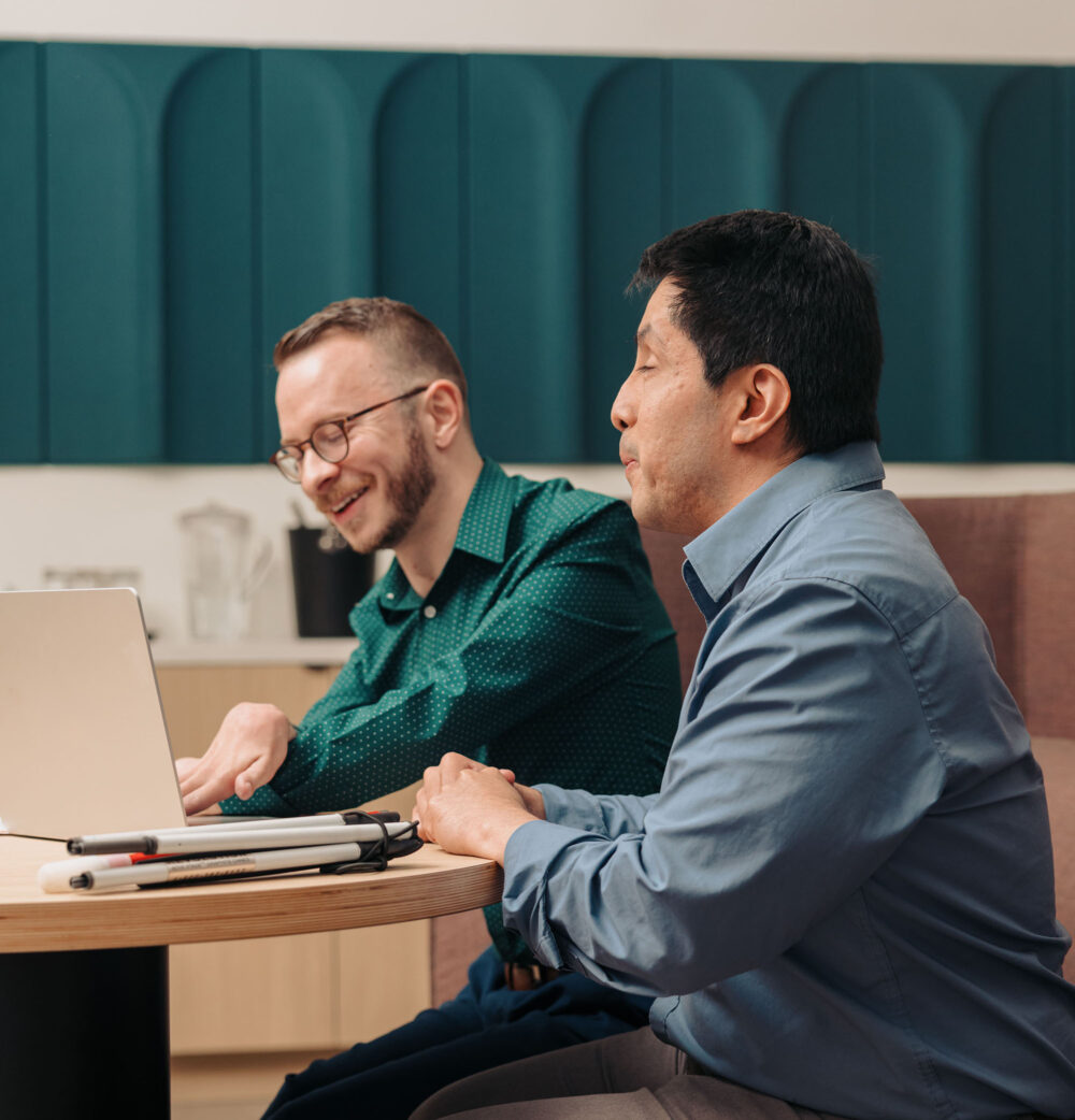 Two colleagues working together on a laptop in an office. One man has his white cane folded on the desk next to the laptop.